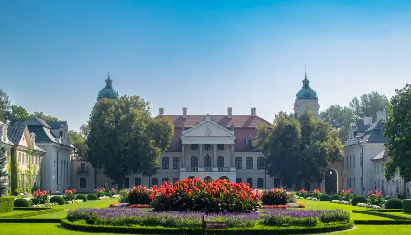 Pałac Zamoyskich w otoczeniu zieleni i kwitnących rabat. Majestatyczna budowla z kolumnowym portykiem i kopułami.
