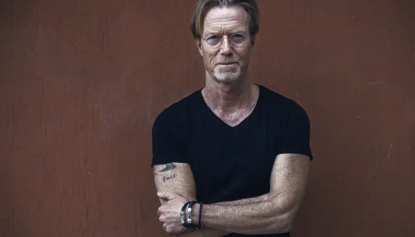 Kommissarie Ewert Grens, en man med intensiv blick och tatuering på armen, står med armarna i kors mot en rödbrun vägg.