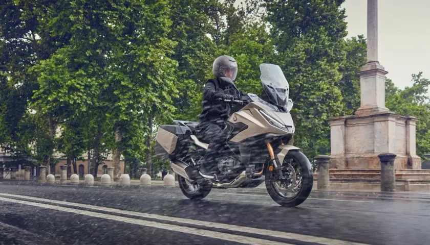 Motociclista en moto beige con alforjas, rodando bajo la lluvia. ¿Cuántos kilómetros dura una moto 125? ¡Esta parece lista para un largo viaje!