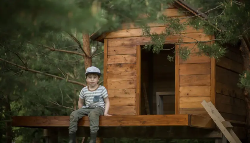 Chłopiec w czapce siedzi na tarasie domku na drzewie. To inspiracja, jak zbudować domek na drzewie dla dzieci.
