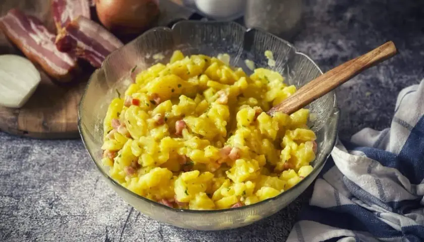 Schüssel mit warmem Kartoffelsalat, garniert mit Speck und Zwiebeln. Ein Holzlöffel liegt darin.