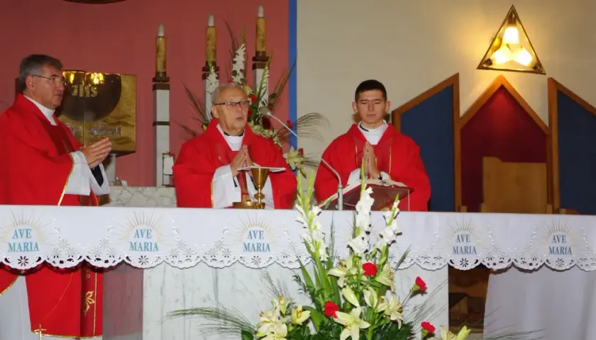 Trzech księży w czerwonych szatach podczas mszy, przed ołtarzem z kwiatami i napisem "Ave Maria".