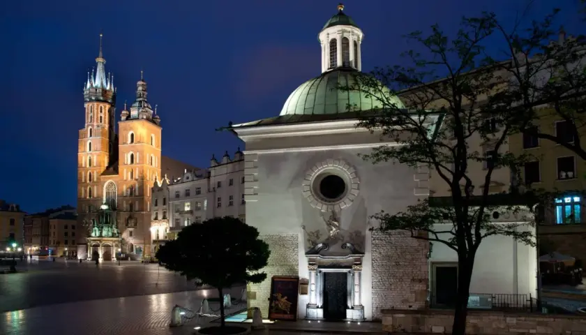 Nocny widok na Rynek Główny w Krakowie. W oddali majestatyczna Bazylika Mariacka, a na pierwszym planie kościół św. Wojciecha z zieloną kopułą.