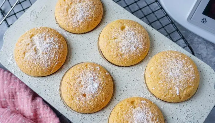 Sechs goldbraune Muffins, mit Puderzucker bestäubt, auf einer hellen Backform. Perfekt für den Thermomix!