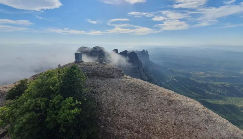 Cumbre rocosa con un monolito en Sant Jeroni, Montserrat. Nubes y montañas se extienden hasta el horizonte.