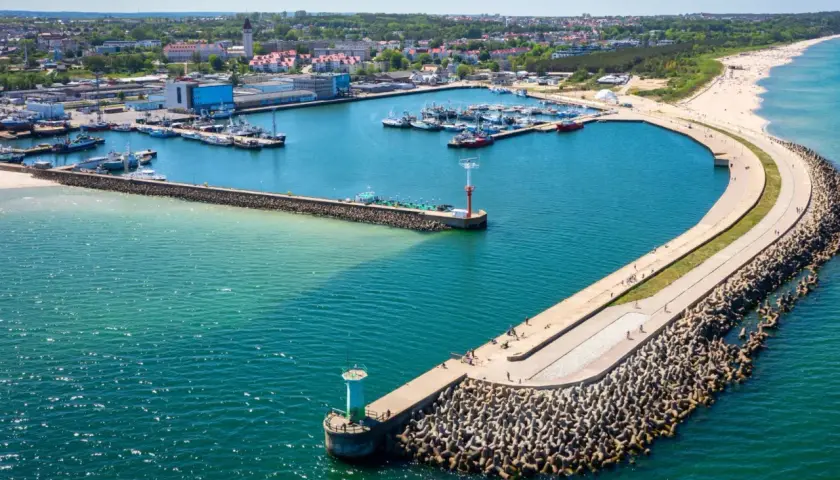 Port w Władysławowie z licznymi łodziami i falochronem, na którym widać spacerujących ludzi. W oddali plaża i zabudowania.