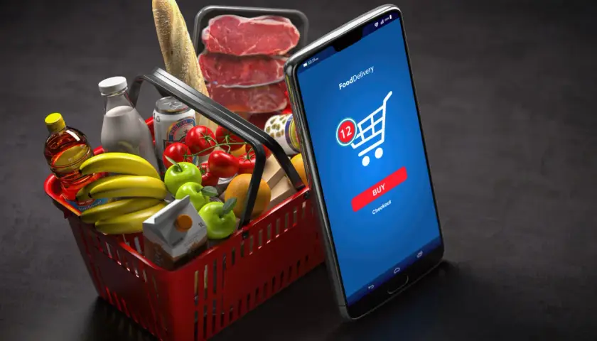 Koszyk z zakupami i smartfon z aplikacją do zamawiania jedzenia. M-commerce ułatwia zakupy spożywcze.