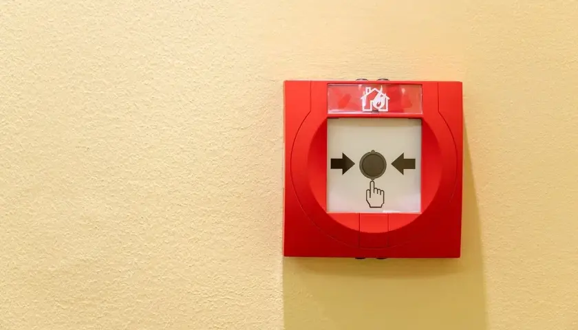 Déclencheur d'alarme incendie rouge sur mur jaune. Une main stylisée pointe le bouton, prêt à signaler une fausse alarme incendie.