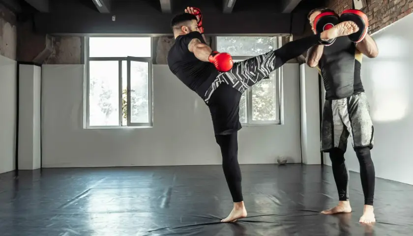 Trener łapie kopnięcie low kick. Dwóch mężczyzn w rękawicach treningowych ćwiczy sztuki walki na sali.