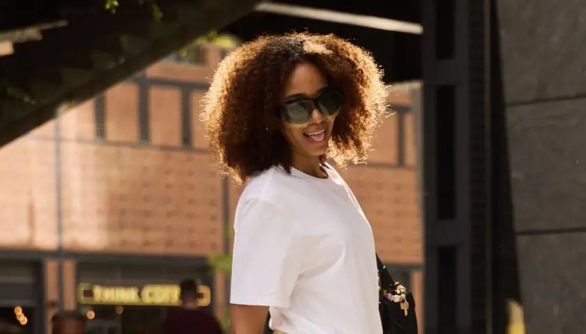 Frau mit lockigem Haar und Sonnenbrille trägt ein weißes T-Shirt.