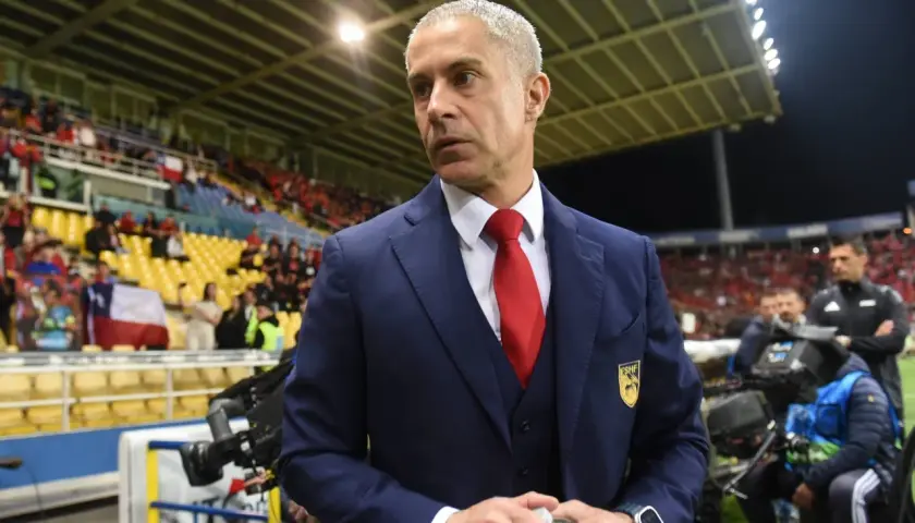 Sylvinho w granatowym garniturze i czerwonym krawacie, z emblematem na piersi, stoi na tle trybun stadionu.