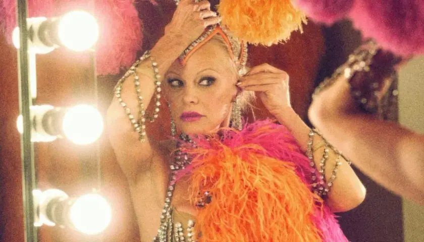 Pamela Anderson w kolorowym piórkowym stroju, przygotowująca się do występu. Jej styl przypomina kreacje z jej filmów.