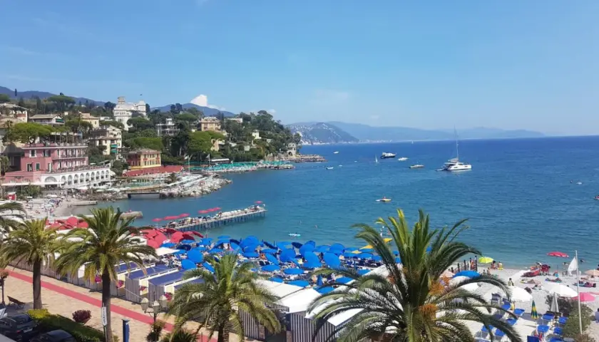 Plage animée à Santa Margherita Ligure, Italie, avec parasols bleus, cabines rayées, palmiers et bateaux sur la mer bleue.