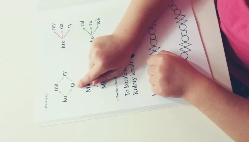 Dziecko ćwiczy czytanie, wskazując palcem na litery alfabetu i sylaby w książce. Obok widnieje wzór z figur geometrycznych.