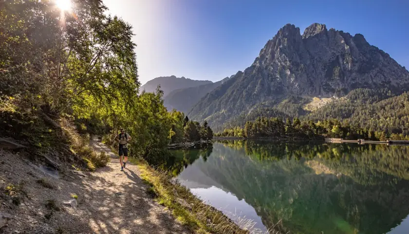 Senderista camina por el sendero junto al estany de Basturs, reflejando la majestuosa montaña y el bosque en sus aguas tranquilas.