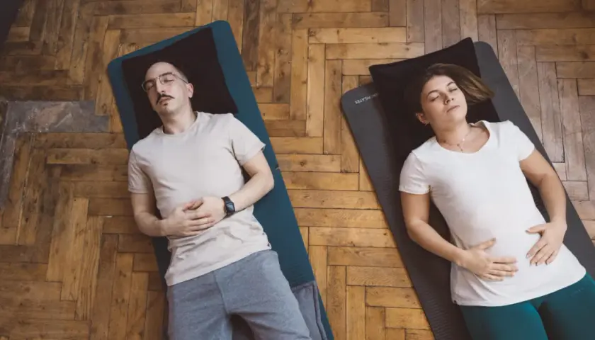 Un couple pratique la respiration profonde technique sur des tapis de yoga. Ils sont allongés sur le dos, les yeux fermés, les mains sur le ventre.