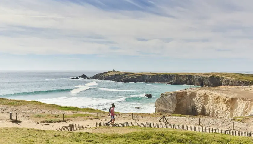 Deux personnes marchent sur un sentier côtier, découvrant les plus beaux endroits de Quiberon. Falaises dorées, mer turquoise et ciel bleu.