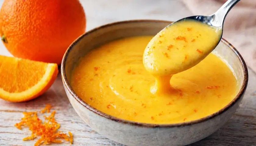 Une cuillère dégouline d'une sauce maltaise onctueuse, parsemée de zestes d'orange. Une orange entière et une tranche sont à proximité.