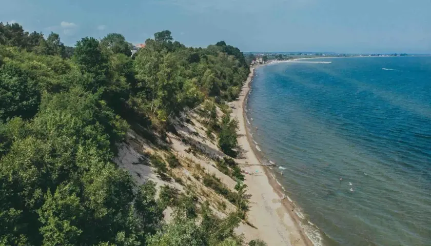 Mechelinki klif porośnięty drzewami, z piaszczystą plażą i turkusowym morzem. W oddali widać zabudowania i falochron.
