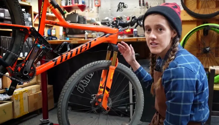 A woman in a blue plaid shirt and beanie explains bike maintenance, pointing to the orange frame and front suspension of a mountain bike.
