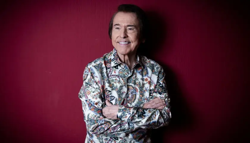 Raphael, sonriente y con los brazos cruzados, posa ante un fondo rojo. Su camisa estampada evoca la energía de sus conciertos.