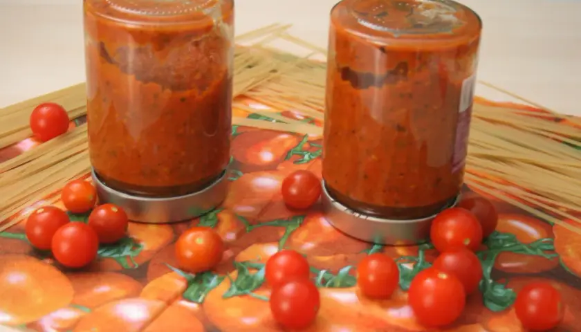 Zwei Gläser mit hausgemachtem tomaten sugo stehen neben Spaghetti. Frische Kirschtomaten zieren die Serviette mit Tomatenmuster.