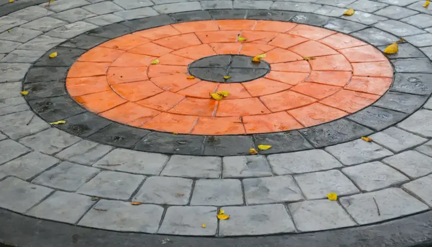 Kreisrunder Weg aus grau und orangefarbenen Pflastersteinen, die wie ein Ziel aussehen. Einige gelbe Blätter liegen darauf.