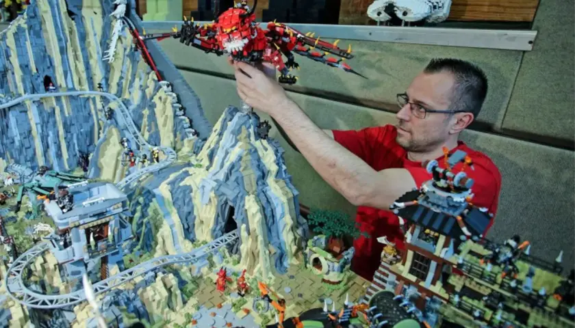 Mann mit Brille platziert roten Drachen in einem riesigen Lego-Set mit den meisten Teilen, das eine Berglandschaft mit Achterbahn und Gebäuden zeigt.