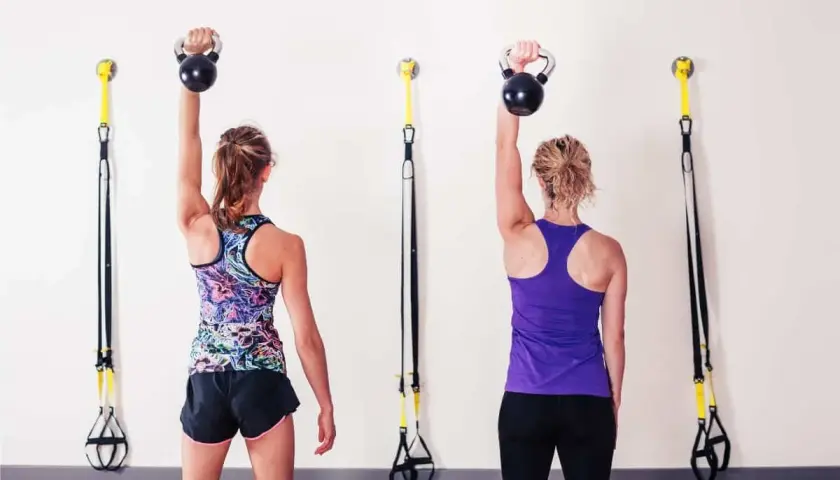 Dwie kobiety wykonują rzs ćwiczenia z kettlebellami. W tle wiszą taśmy TRX.