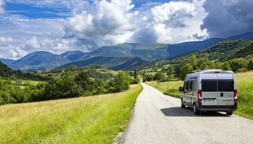 Un camping-car Elios sillonne une route sinueuse à travers les paysages verdoyants du Vercors, sous un ciel bleu parsemé de nuages.