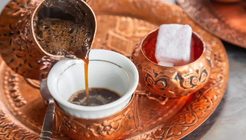 Frisch aufgebrühter, bester türkischer Kaffee fließt aus einer Kupferkanne in eine kleine Tasse. Daneben liegen süße Loukoum-Würfel.