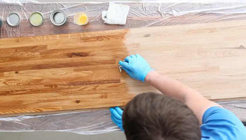 Mano con guanto blu applica una finitura su legno, creando una patina. Diverse lattine di vernice e un panno sono visibili.