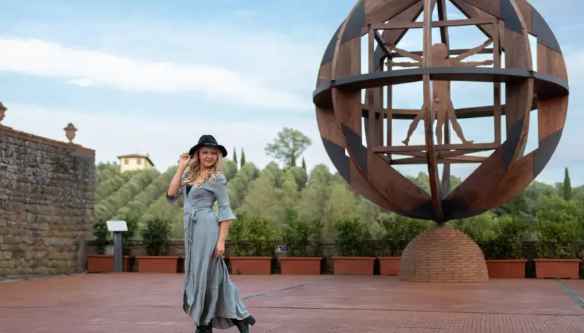 Donna con cappello nero e abito lungo in un'area panoramica del vinci centro storico, con la scultura dell'Uomo Vitruviano sullo sfondo.