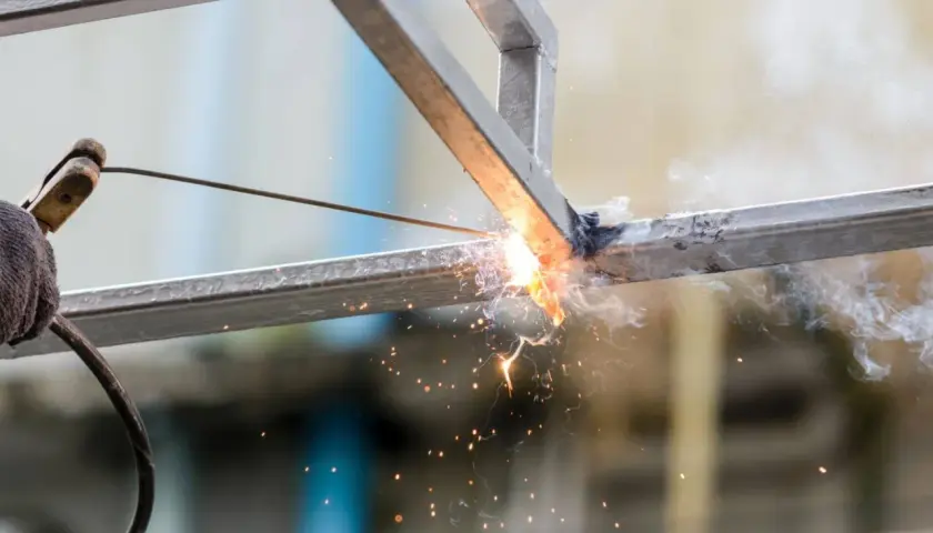 Ręka w rękawicy trzyma elektrodę, wykonując spawanie łukowe metalowych profili. Iskry i dym unoszą się w powietrzu.