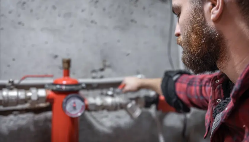 Uomo con barba controlla un manometro su un tubo rosso. Sento rumore nei tubi dell'acqua, forse un problema da risolvere.