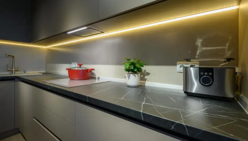 Nowoczesna kuchnia z eleganckim oświetleniem LED nad blatem. Czerwony garnek i toster dodają koloru. Idealne rozwiązanie, jak oświetlić kuchnię.