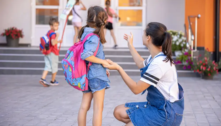 Kobieta klęczy przed dziewczynką z plecakiem, rozmawiając z nią przed szkołą.
