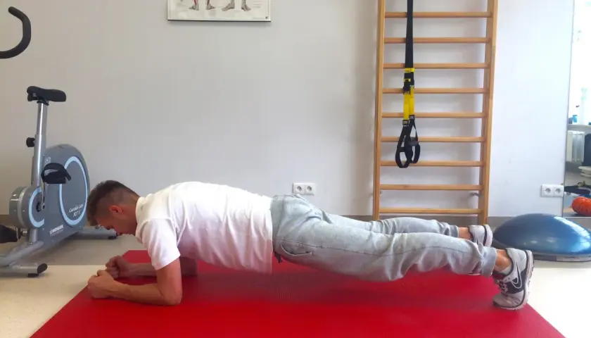 Mężczyzna wykonuje plank, wzmacniając mięśnie core. Obok rower stacjonarny i drabinka gimnastyczna.