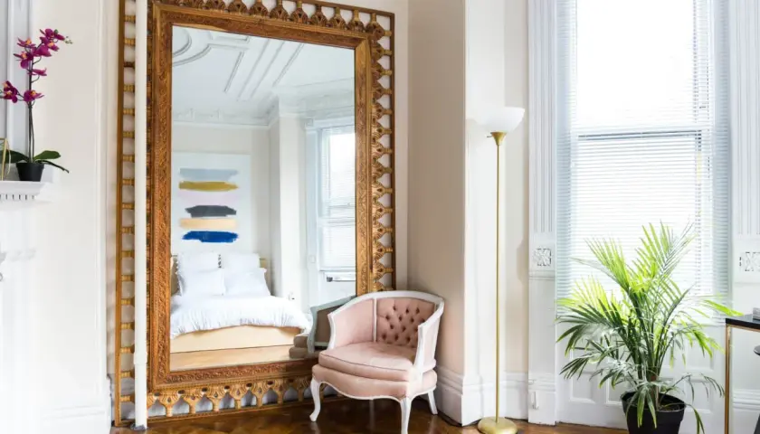 A grand pier mirror with an ornate gold frame reflects a bed and abstract art. A pink velvet chair sits beside it.