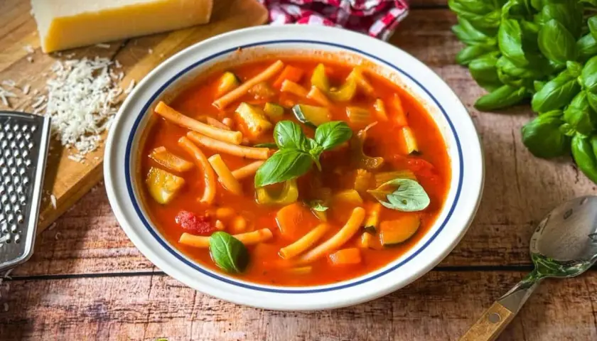 Herzhaftes rezept minestrone original mit viel Gemüse und Pasta, garniert mit frischem Basilikum. Daneben Parmesan und ein Löffel.