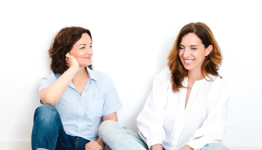 Dos mujeres sentadas, una con camisa azul y jeans, la otra con blusa blanca. Parecen compartir un momento laconicum, de pocas palabras pero profunda conexión.