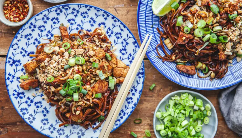 Två tallrikar med läcker thai kyckling med nudlar, toppad med jordnötter och salladslök. Serveras med limeklyftor och chiliflakes.