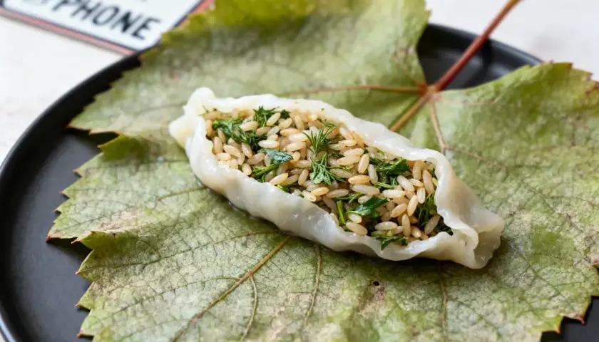 Une feuille de vigne farcie, garnie de riz et d'herbes fraîches, repose sur une grande feuille de vigne verte sur une assiette noire.
