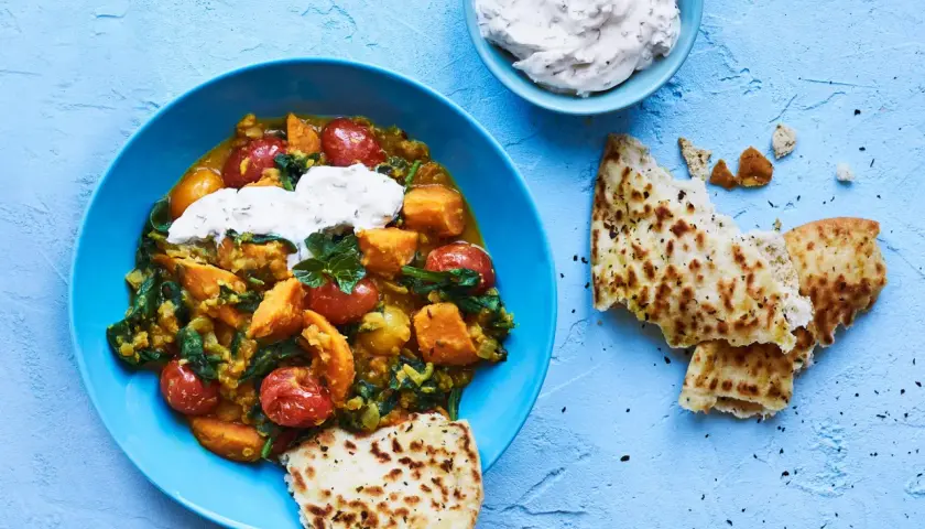 En blå skål med en värmande daal med sötpotatis, spenat och tomater, toppad med gräddfil. Serveras med naanbröd.