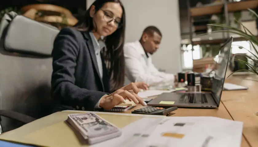 Kobieta w okularach liczy na kalkulatorze, obok stos banknotów i laptop.