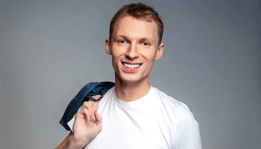 Uśmiechnięty Igor Herbut w białej koszulce z dżinsową kurtką na ramieniu.