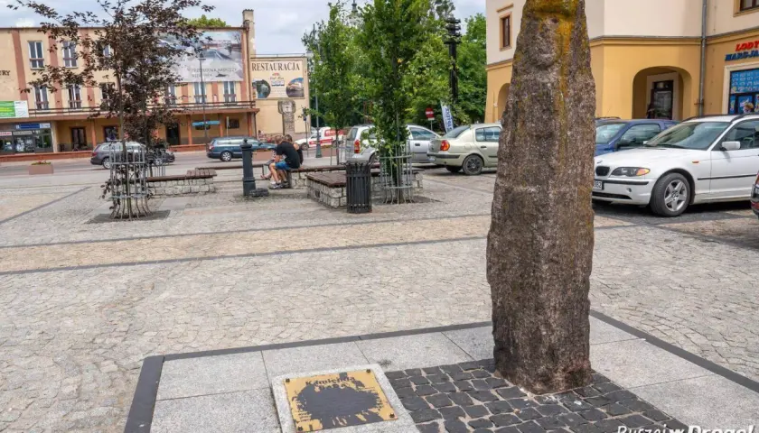 Kamienny pomnik i tablica z napisem "Kamienna" to ciekawe atrakcje na rynku. Obok ławki i drzewa, w tle budynki i samochody.