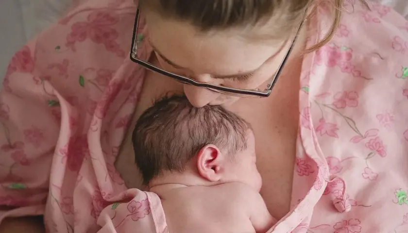 Mama całuje główkę noworodka, otulając go w różowy materiał. To piękny moment kangurowania, budowania więzi.