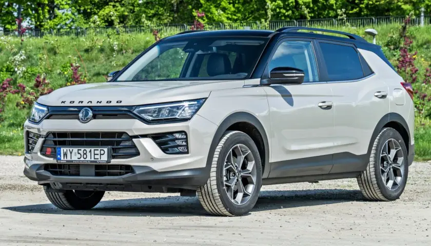 Jasnoszary SsangYong Korando z czarnym dachem stoi na drodze gruntowej, otoczony zielenią.