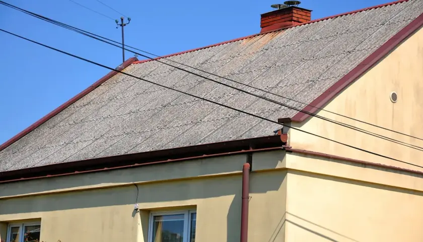 Dach pokryty falistym eternitem, z ceglanym kominem i liniami energetycznymi przecinającymi błękitne niebo.